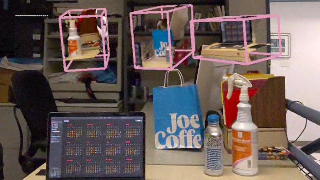 Headset passthrough view of a lab desk. Gaussian-splatted proxies of a spray bottle, a bag and a laptop float in pink wireframe boxes above the real bag, bottles and laptop on the desk.